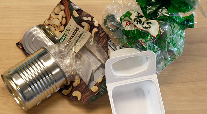 Verschiedene Verkaufsverpackungen, unter anderem eine Nussverpackung, Konservendose und Joghurt liegen auf einem Tisch.