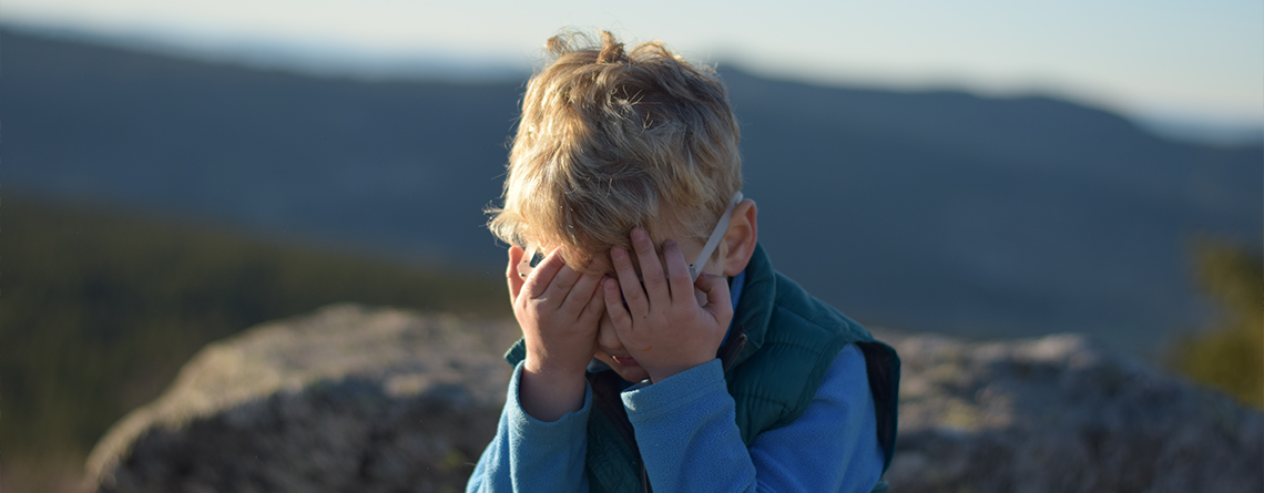 Ein Kind in der Natur hält sich mit beiden Händen die Augen zu, der Kopf ist gesenkt