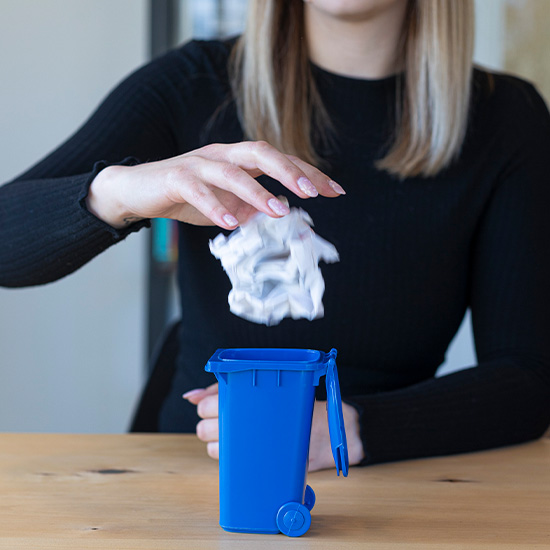 Frau schmeißt ein zerknülltes Blatt Papier in eine blaue Papiertonne im Miniaturformat.